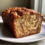 A close-up of a freshly baked, moist apple bread loaf showing a rich brown crust and visible chunks of nuts inside.