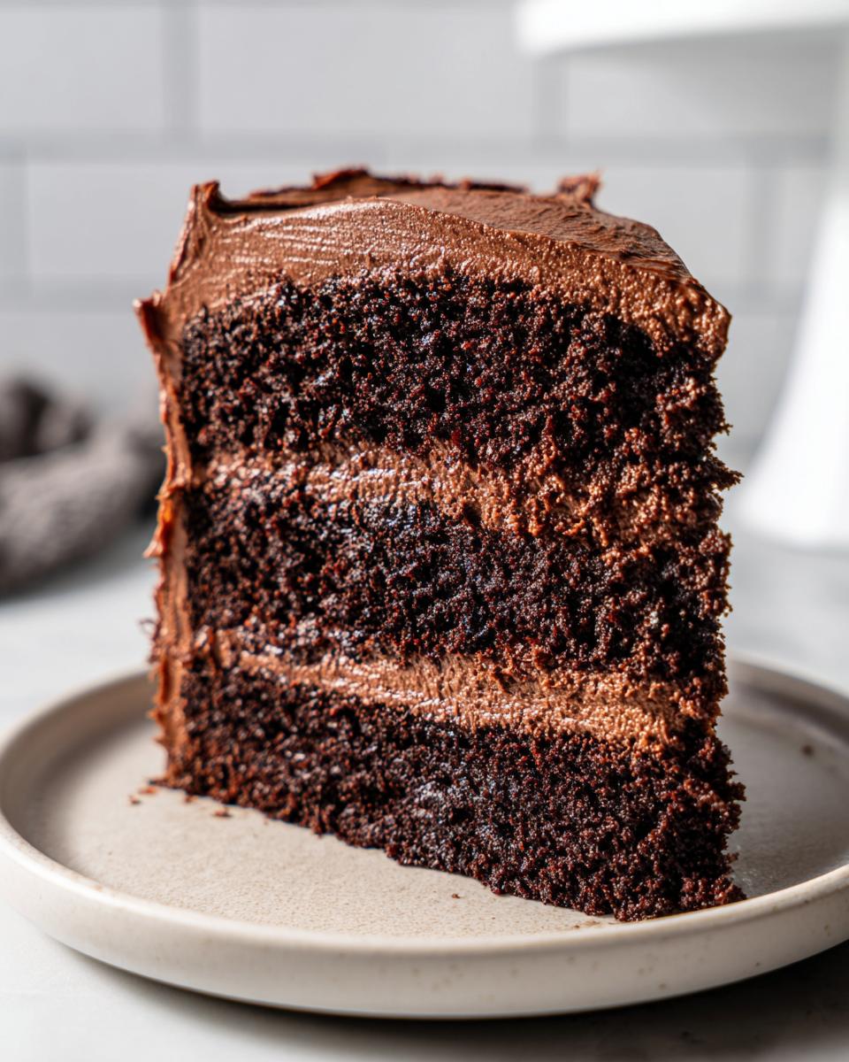 A tall, dark slice of moist chocolate cake showing three layers of cake and two layers of chocolate frosting.