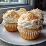 Close-up of four moist coconut cupcakes topped with white frosting and toasted coconut flakes.