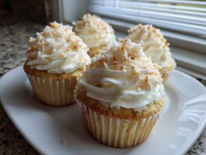 Four moist coconut cupcakes topped with white frosting and toasted coconut flakes, arranged on a white plate.