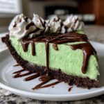 A decadent slice of no-bake grasshopper pie featuring a bright green filling, dark chocolate crust, and chocolate syrup drizzle.