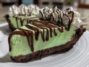 A close-up of a slice of bright green, no-bake grasshopper pie with a dark chocolate crust, topped with whipped cream and chocolate syrup.
