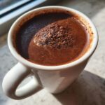 Close-up of a rich, dark brown beverage made from hot cocoa mix in a white mug, topped with foam.