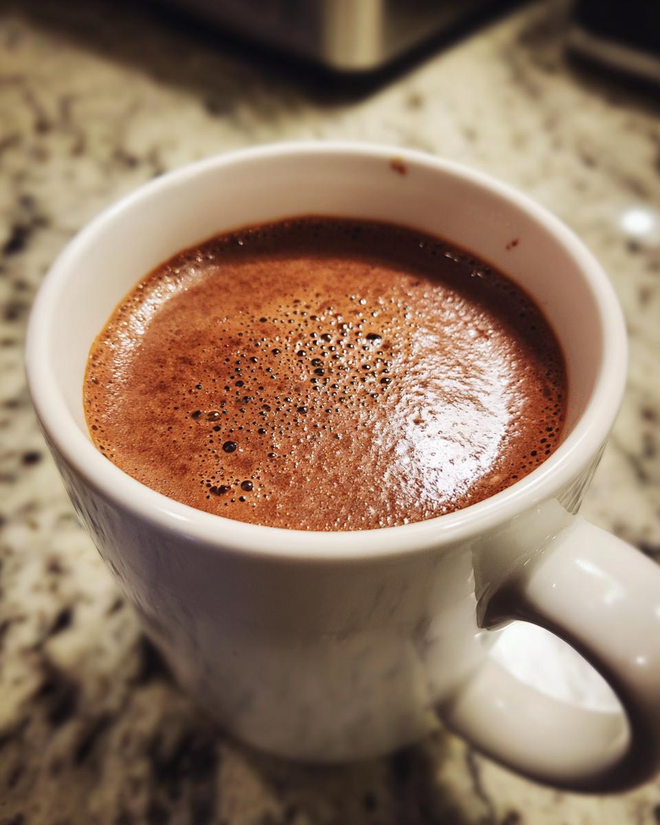 Close-up of rich, frothy hot cocoa mix in a white ceramic mug sitting on a granite countertop.