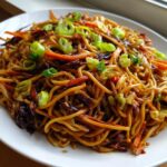 Close-up of a white plate piled high with savory fried noodles, mixed with shredded carrots and cabbage, topped with fresh green onions.