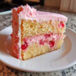 A close-up of a slice of layered cherry cake featuring yellow sponge, pink cherry filling, and pink frosting.