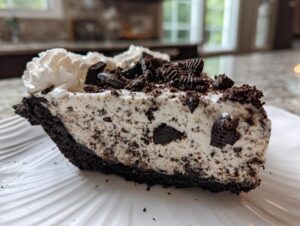 Close-up of a decadent slice of ice cream pie featuring a dark cookie crust, creamy filling studded with cookie pieces, and topped with whipped cream.