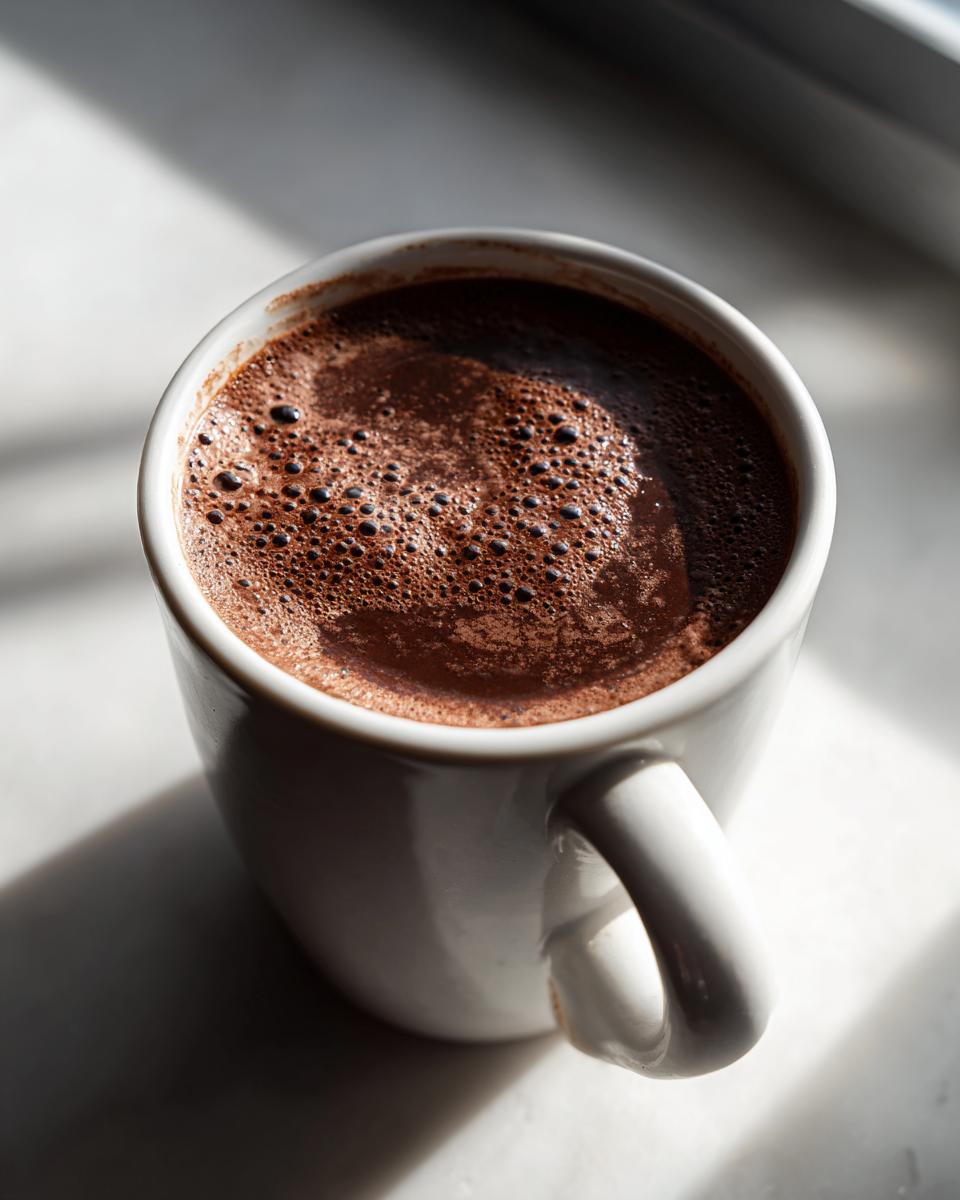 Close-up of a white mug filled with rich, frothy hot cocoa mix, highlighted by strong sunlight.