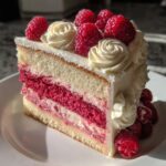 A perfect slice of stunning rose cake featuring layers of white sponge, bright pink raspberry filling, and white frosting rosettes topped with fresh raspberries.