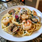 Close-up of a white plate filled with linguine seafood pasta featuring shrimp, clams, scallops, and herbs.
