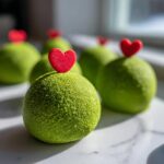 Close-up of vibrant green Grinch Oreo balls topped with small red heart sprinkles on a white marble surface.