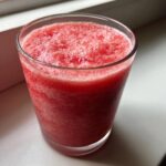 Close-up of a vibrant red watermelon smoothie with a frothy top in a clear glass.