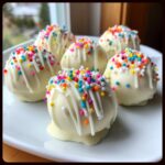 Six white chocolate coated sugar cookie truffles topped with colorful sprinkles and white chocolate drizzle.