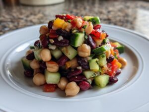 A vibrant mound of fresh bean salad featuring chickpeas, kidney beans, cucumber, and peppers on a white plate.