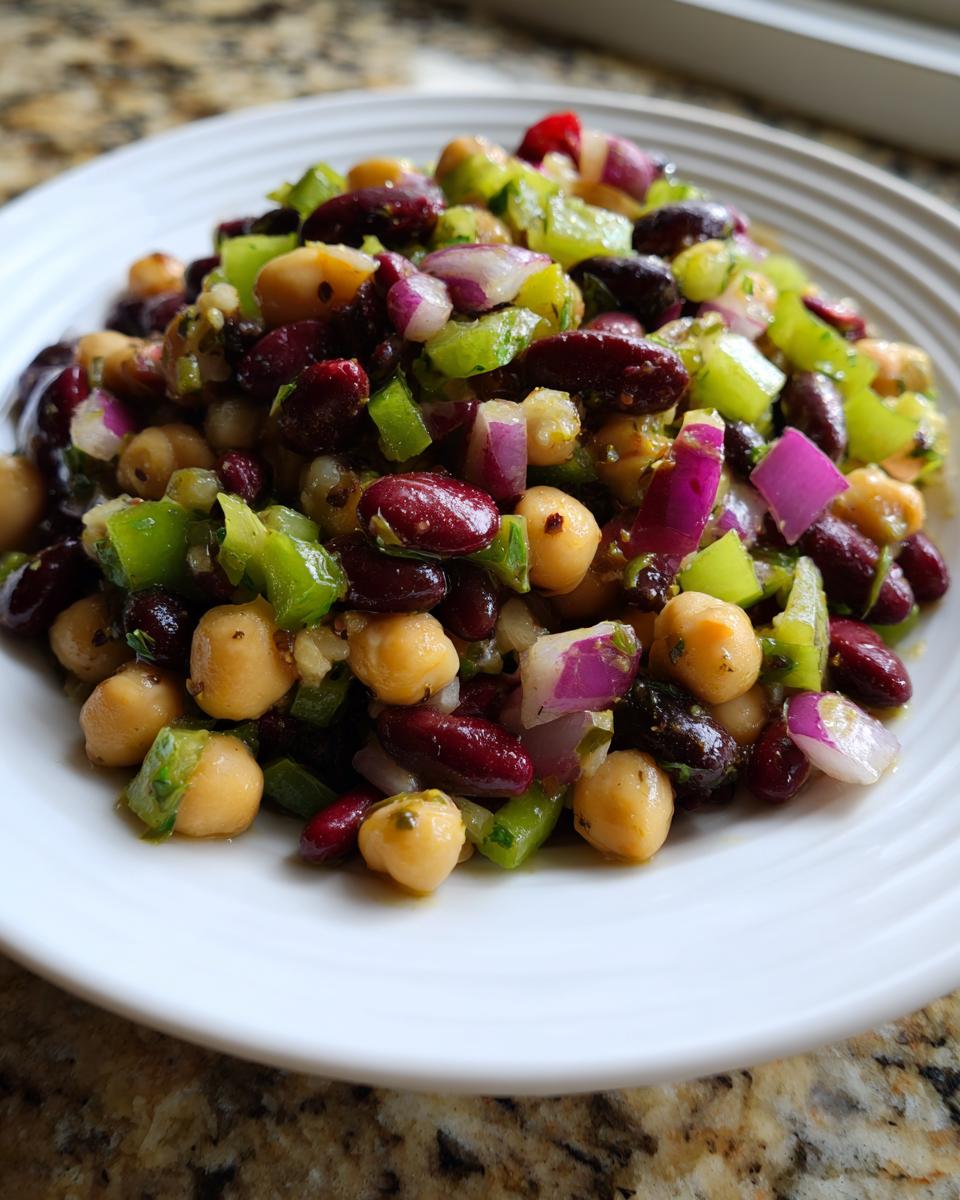 Close-up of a serving of dense bean salad featuring kidney beans, chickpeas, green peppers, and red onion on a white plate.