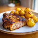 A close-up of a perfectly seasoned baked pork chop next to a serving of golden roasted potatoes.