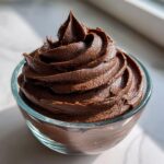 A close-up of rich, dark chocolate buttercream frosting swirled high in a clear glass bowl.