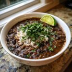 A close-up of a bowl of rich black bean and rice soup, topped with white rice, cilantro, and a lime wedge.