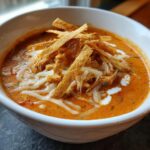 A close-up bowl of vibrant orange cheesy chicken tortilla soup topped with shredded cheese, chicken, and crispy tortilla strips.