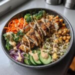 A vibrant chicken buddha bowl featuring sliced seasoned chicken, quinoa, broccoli, carrots, cucumbers, and chickpeas, drizzled with sauce.