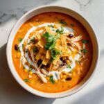 Overhead view of a bowl of rich, orange chicken tortilla soup topped with shredded chicken, beans, cream, and tortilla strips.