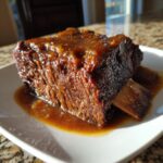 A single, tender piece of cider braised short ribs covered in a thick, glossy brown sauce on a white plate.