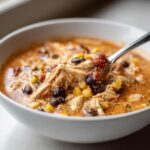A spoonful of creamy 7 can chicken taco soup, showing shredded chicken, black beans, and corn, being lifted from a white bowl.