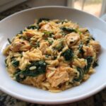 Close-up of creamy orzo pasta mixed with chunks of chicken and wilted spinach served in a white bowl.