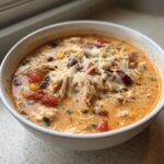 A close-up of a white bowl filled with creamy chicken taco soup, featuring shredded chicken, beans, corn, tomatoes, and melted cheese.