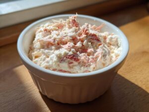 A small white bowl filled with creamy, pink and white imitation crab dip recipe mixture, sitting on a wooden surface.