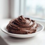 A swirl of rich, dark chocolate buttercream frosting resting on a small white plate near a bright window.