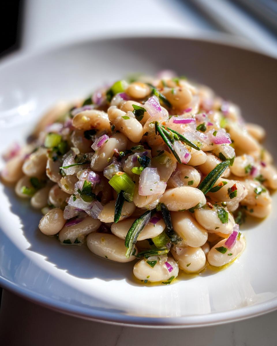 A close-up of a vibrant white bean salad featuring cannellini beans mixed with chopped red onion, celery, and fresh rosemary.