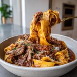 A fork lifts wide ribbons of pasta coated in rich braised short rib pasta sauce from a bowl.