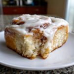 A close-up of a single, soft cinnamon roll recipe serving, dripping with white cream cheese frosting.