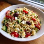 Close-up of a bright orzo pasta salad recipe mixed with cherry tomatoes, cucumber, feta, and Kalamata olives.