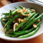 Close-up of a white bowl filled with vibrant green bean salad topped with caramelized onions and sliced almonds.