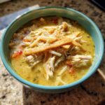 A close-up of a bowl of creamy green chile chicken enchilada soup topped with shredded chicken, cheese, and tortilla strips.