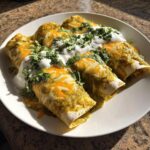 Three rolled green chile chicken enchiladas corn tortillas covered in green sauce, melted cheese, sour cream, and cilantro.
