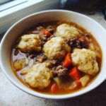 A close-up of a bowl filled with rich ground beef and dumpling soup, featuring tender dumplings, ground beef, carrots, and onions.