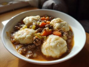 A close-up of a white bowl filled with savory ground beef and dumplings crockpot stew, featuring carrots and celery.