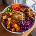 Close-up of a vibrant buddha bowl featuring crispy tofu, quinoa, peanut sauce, shredded carrots, edamame, and red cabbage.