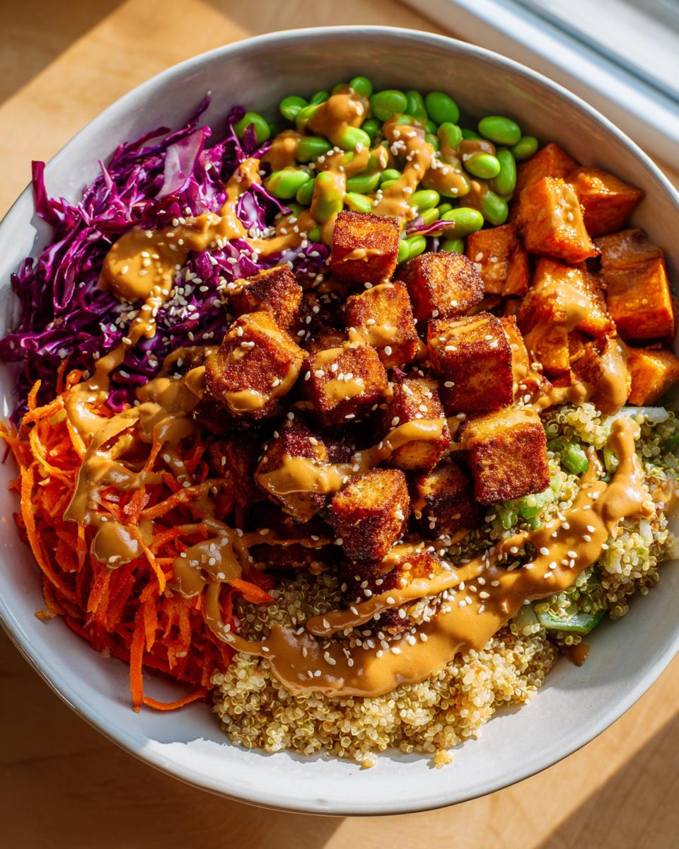 Overhead view of a colorful buddha bowl featuring crispy tofu, quinoa, shredded carrots, red cabbage, edamame, and peanut sauce.