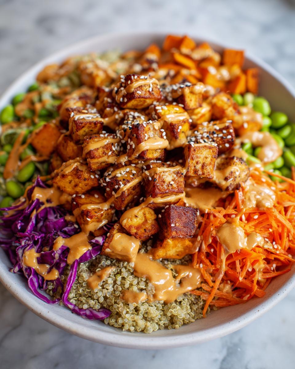 Close-up of a vibrant buddha bowl featuring crispy tofu cubes, quinoa, shredded carrots, red cabbage, and edamame, drizzled with peanut sauce.