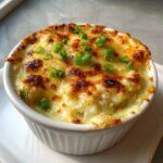 Close-up of hot baked crab dip in a white ramekin, topped with melted, browned cheese and fresh green onions.