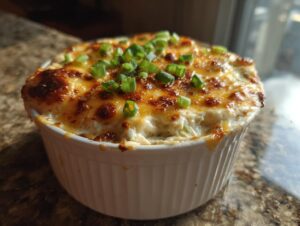 Close-up of a hot baked crab dip in a white ramekin, topped with bubbly, browned cheese and sliced green onions.