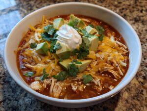 A close-up of a white bowl filled with rich, red keto chicken tortilla soup, topped with shredded cheese, avocado, sour cream, and cilantro.