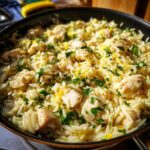 Close-up of bright lemon chicken orzo pasta with chicken pieces and fresh parsley in a dark skillet.