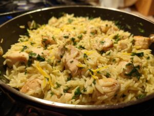 Close-up of finished lemon chicken orzo pasta with chunks of chicken and fresh parsley in a dark skillet.