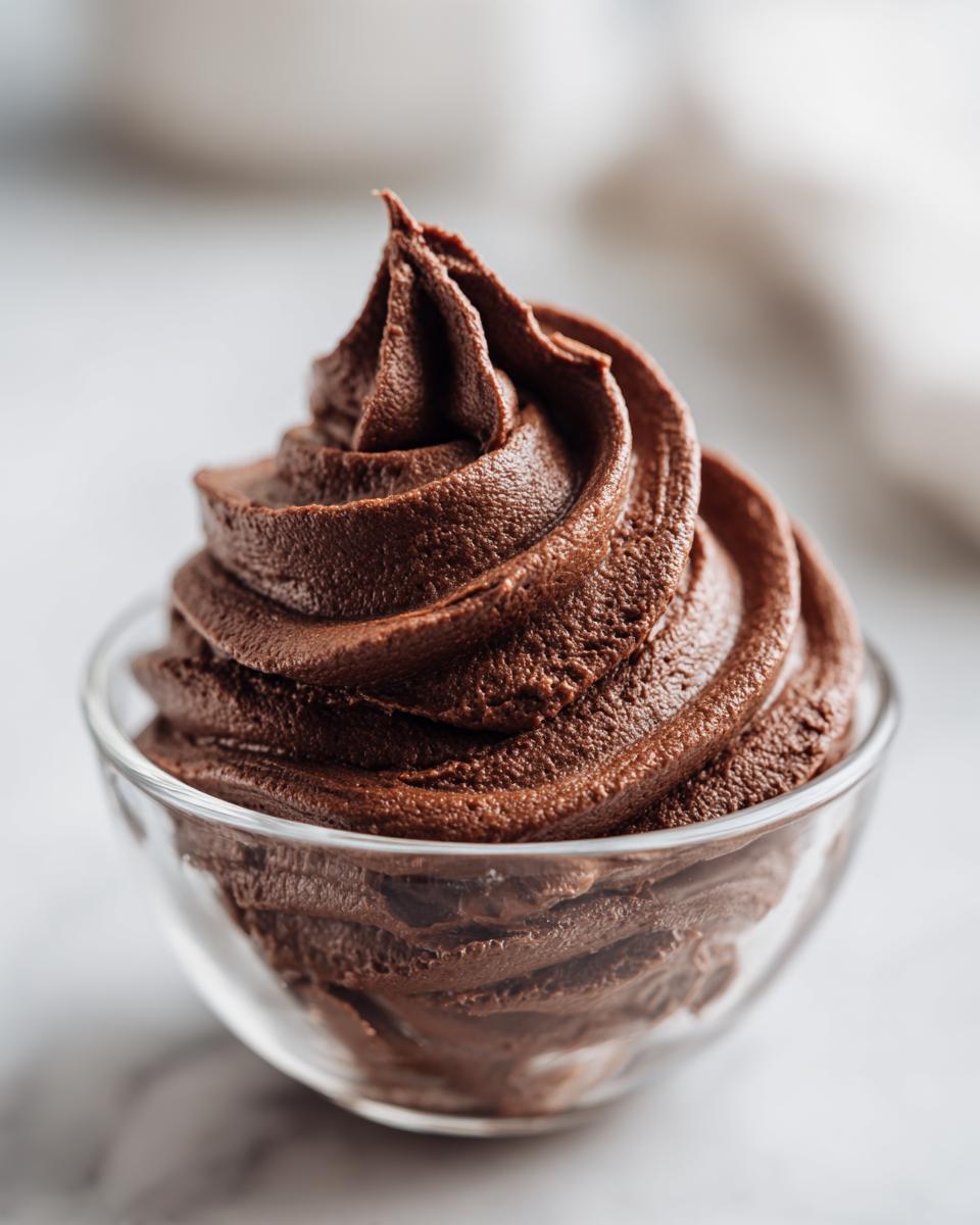 Close-up of rich, dark chocolate buttercream frosting piped into a perfect swirl in a small glass bowl.
