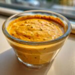 Close-up of a vibrant, orange-hued southwest salad dressing with visible specks of herbs and red pepper flakes in a clear glass dish.
